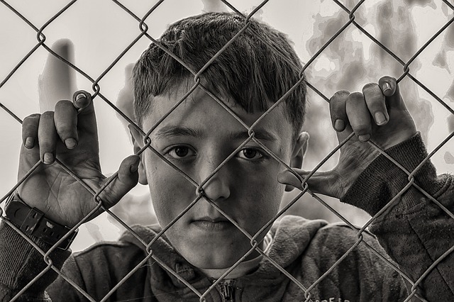 A black and white picture of a young boy looking out through a chain link fence.