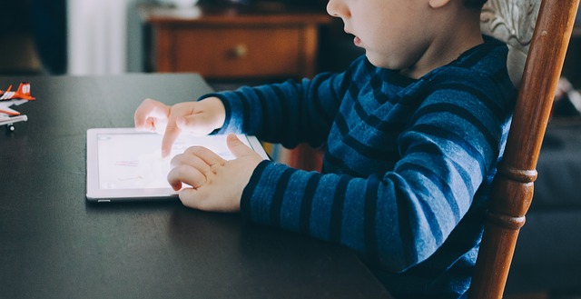 A child sitting at a table as they look at a tablet screen and touch the screen to interact with a game.