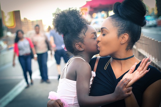 A young woman kissing a little girl, while a couple stand in the background, out of focus.