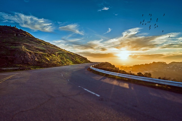 A winding road that curves away into a bright sunrise, with wispy clouds and birds in the sky.