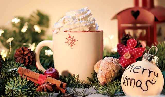 A cup of hot chocolate with shipped cream, nestled into a snowy pine bough with cinnamon sticks, lights, and ornaments.