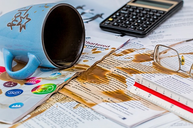 A tipped over cup of coffee, with coffee spilled onto piles of printed paper.