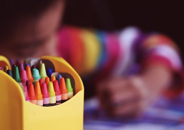 A close up of an open box of crayons, with an out-of-focus young child coloring at a desk in the background.