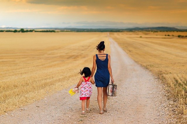 A mother and child walking away down a road through fields, hand in hand with a suitcase.