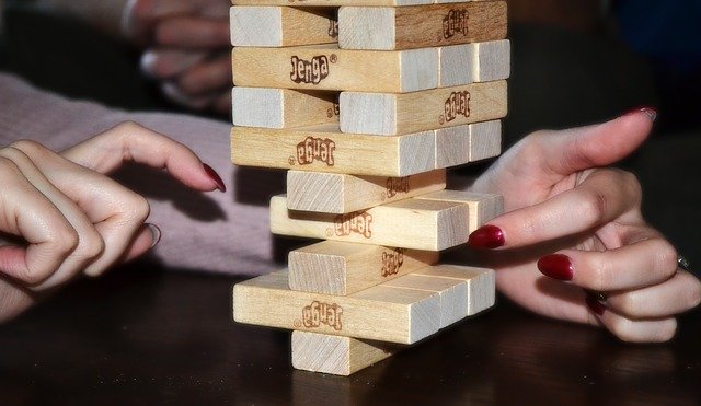 A jenga tower with several pieces missing from the bottom, and a woman's hand slowly sliding another piece out of the bottom, making the tower more likely to fall.