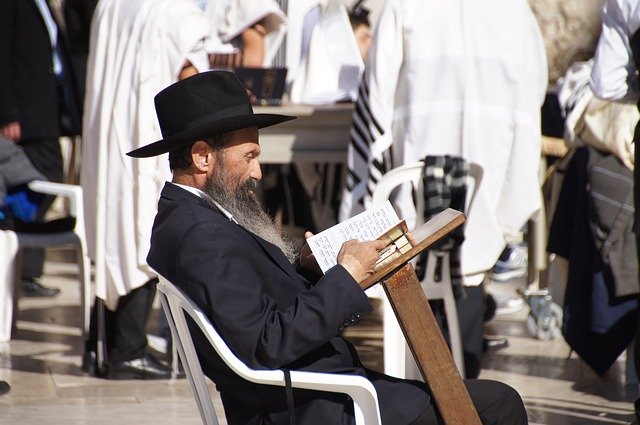 A man in traditional Judaic clothing, sitting in a public park, reading the Torah.