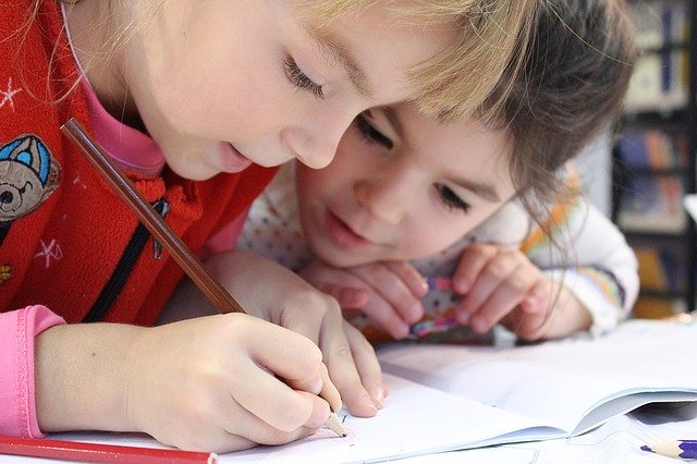 Two little girls at school, leaning over a work book and solving a math problem together.