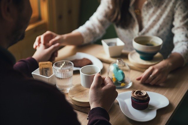 A man and woman sitting at a table drinking coffee and eating cupcakes and holding hands.