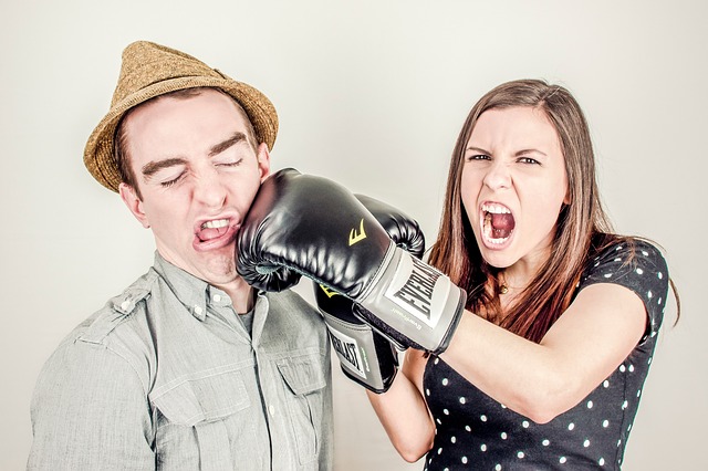 A man and woman fighting. She is wearing boxing gloves and punching the man in the face.