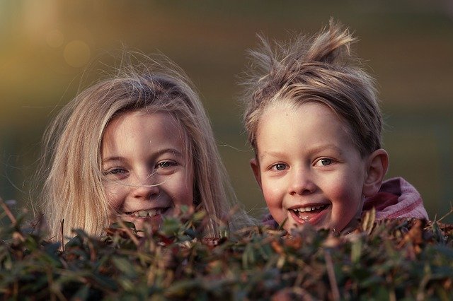 A young boy and girl peeking over a hedge and laughing.