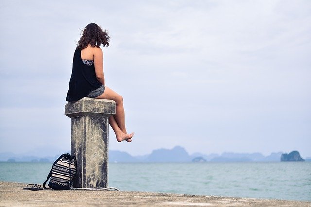 A woman sitting alone on a pillar by the beach, looking out at the sea. There is a bag on the ground at her feet.