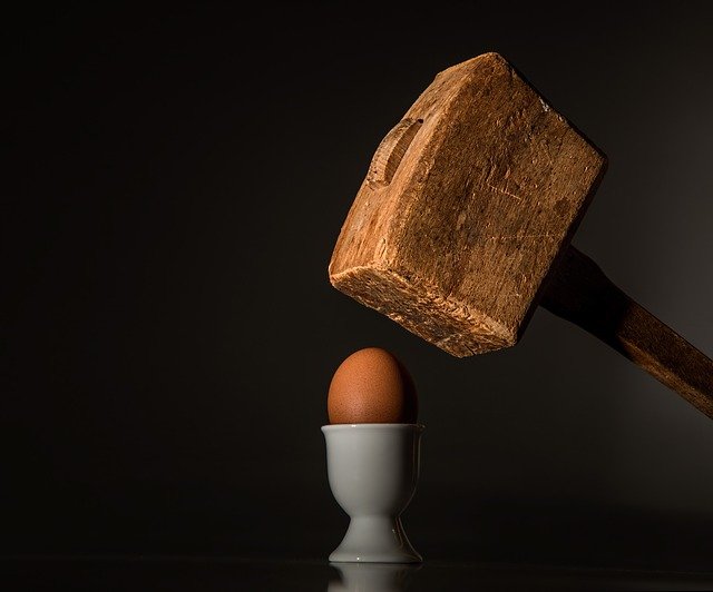 A large wooden mallet coming down to crush a delicate egg in an egg cup.