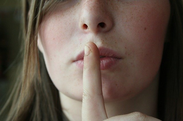 A close up of a woman's face, as she holds a finger to her lips to signify that she has a secret.