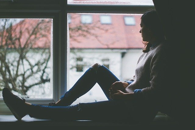A woman sitting in a darkened room, looking sadly out of a window.