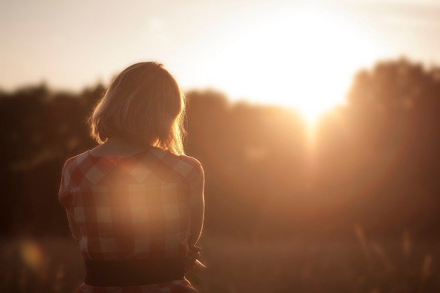 A lonely looking woman standing with her back to the camera, watching the sun set.