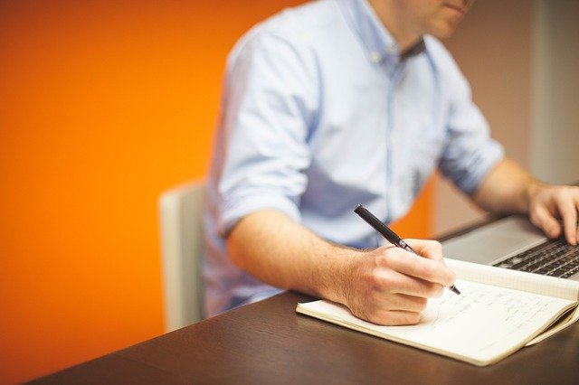 A man in a work shirt sitting at a table making notes in a book while he looks at a laptop screen