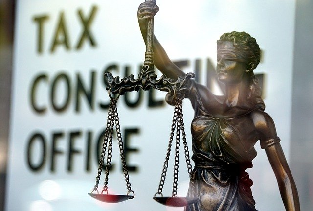 A statue of lady justice, wearing her blindfold and holding up scales, in front of a sign that says "Tax Consulting Office"