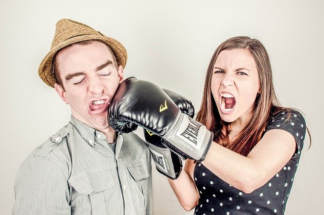 A fighting couple. She is wearing boxing gloves and punching the man in the face while shouting angrily.
