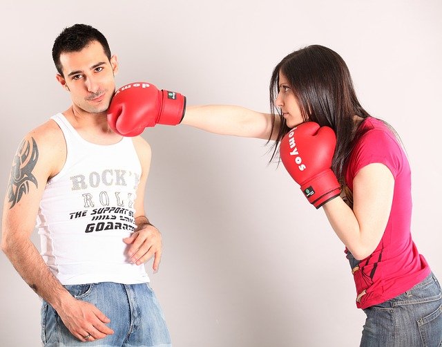 A couple fighting. She is wearing boxing gloves and punching him in the neck.